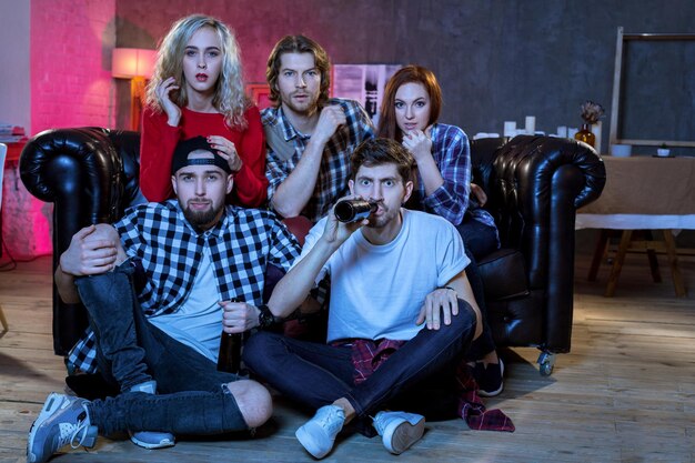 Photo groupe d'amis s'amusant à la maison, regardant un match et s'amusant ensemble, assis dans le canapé du salon la nuit avec un match de championnat à la recherche de pop-corn