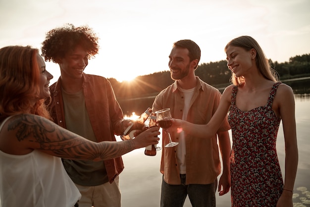 Groupe d'amis s'amusant en pique-nique près d'un lac ou d'une rivière, buvant du vin, de la bière, du cidre trinquant tout en portant un toast. Jeunes souriants faisant la fête à l'extérieur pendant le coucher du soleil dans la campagne.