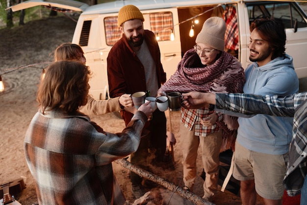 Groupe d'amis tenant des tasses avec du thé chaud et grillant ensemble pendant le pique-nique dans la forêt