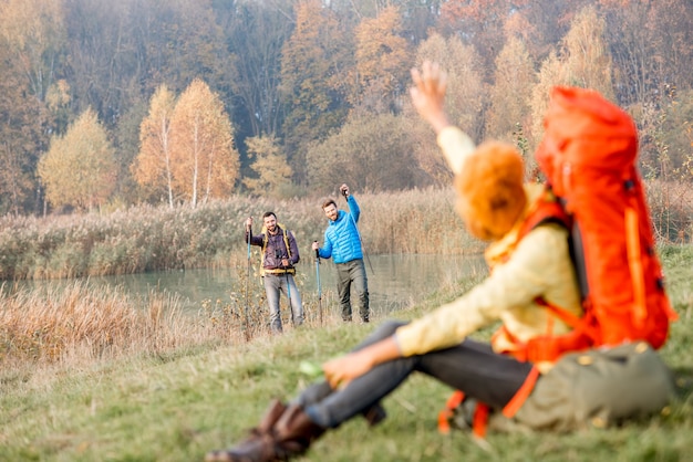Groupe d'amis en vestes colorées avec sacs à dos s'amusant à l'extérieur sur la pelouse verte près du lac et de la forêt