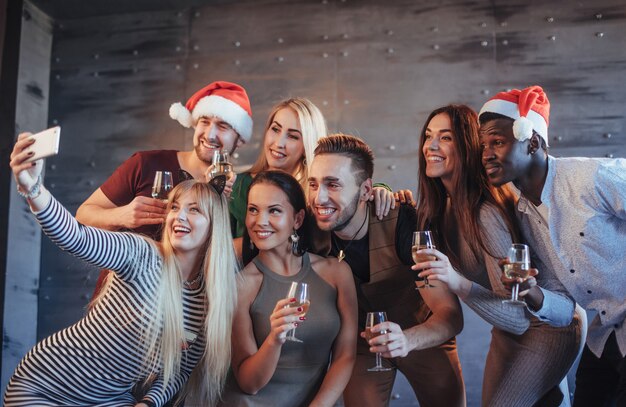 Groupe de beaux jeunes gens faisant du selfie dans la fête du nouvel an, meilleurs amis filles et garçons s'amusant ensemble, posant des gens de style de vie émotionnel Chapeaux santas et verres de champagne dans leurs mains