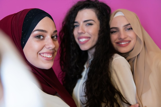 Groupe de belles femmes musulmanes dont deux en robe à la mode avec hijab utilisant un téléphone portable tout en prenant une photo de selfie isolée sur fond rose représentant la technologie de la mode islamique moderne.