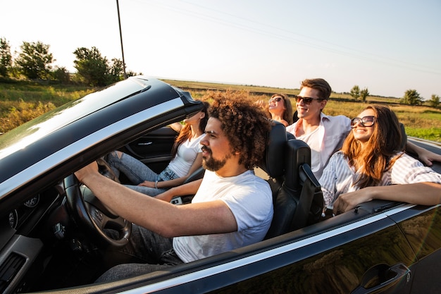 Un groupe de belles jeunes filles et de mecs à lunettes de soleil sourient et roulent dans un cabriolet noir sur la route par une journée ensoleillée. .