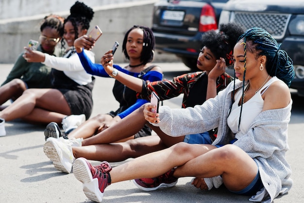 Groupe de cinq femmes afro-américaines assises sur la route ensemble contre une voiture suv sur le parking et faisant du selfie au téléphone