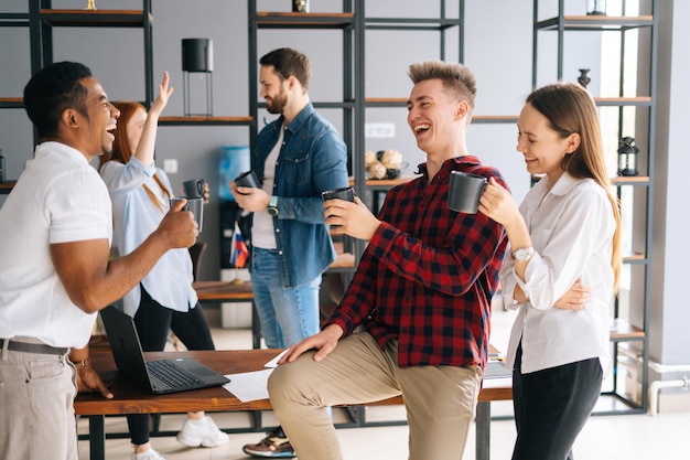 Groupe de cinq jeunes collègues de démarrage multiraciaux qui rient et profitent d'une conversation agréable pendant la pause-café dans un espace de coworking