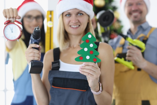 Groupe de contremaîtres réparateurs en chapeaux de père noël tenant une perceuse et un réveil près de l'arbre de noël en gros plan. Concept professionnel de fêtes d'entreprise du nouvel an