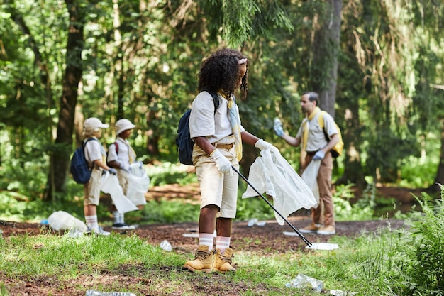 Photo groupe diversifié d'enfants scouts aidant à nettoyer la forêt et tenant des sacs poubelles