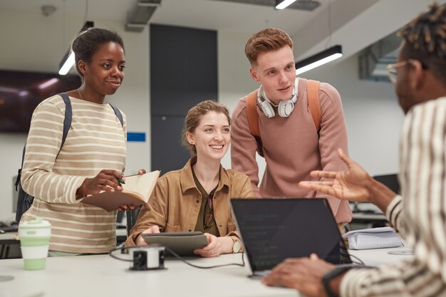 Groupe diversifié d'étudiants discutant du projet au bureau tout en travaillant ensemble à la bibliothèque de l'école et souriant joyeusement