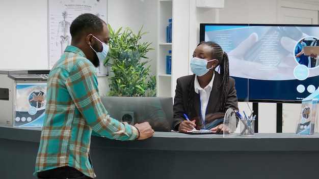 Groupe diversifié de patients avec des masques faciaux assis à la réception de l'hôpital, parlant à la réceptionniste d'un rendez-vous médical. Personnes faisant des consultations pendant l'épidémie de coronavirus.