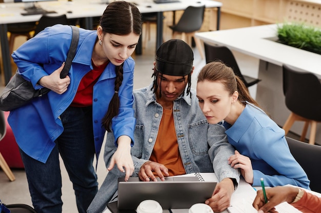 Groupe diversifié de trois étudiants utilisant un ordinateur portable ensemble dans la bibliothèque tout en travaillant sur un projet