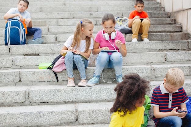 Groupe d'écoliers de différentes nationalités, dans des vêtements colorés assis sur des marches de pierre. Des adolescents qui parlent, écoutent de la musique avec des écouteurs, lisent des livres.