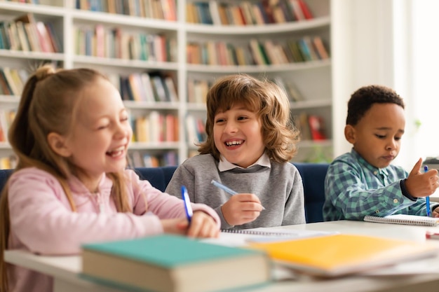 Groupe d'élèves internationaux drôles et divers parlant et riant assis à table avec des cahiers et des livres en classe