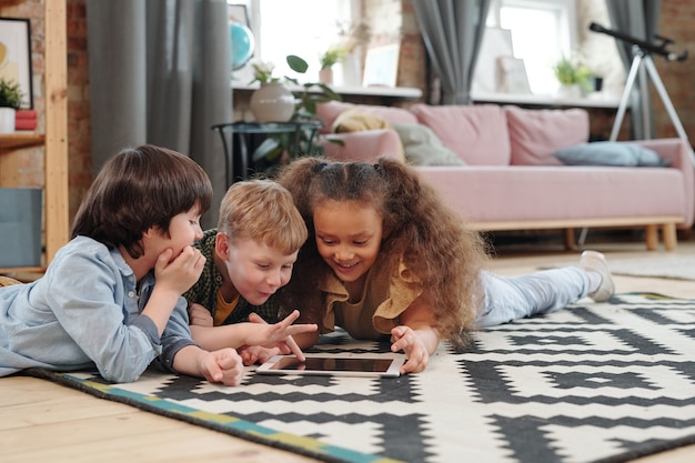 Groupe d'enfants heureux allongés sur le sol et jouant ensemble sur une tablette numérique dans la pièce à la maison