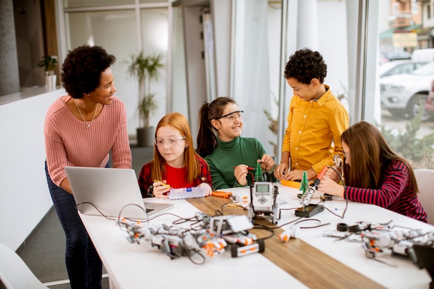 Groupe d'enfants heureux avec leur professeur de sciences afro-américaine avec des jouets électriques de programmation d'ordinateur portable et des robots en classe de robotique