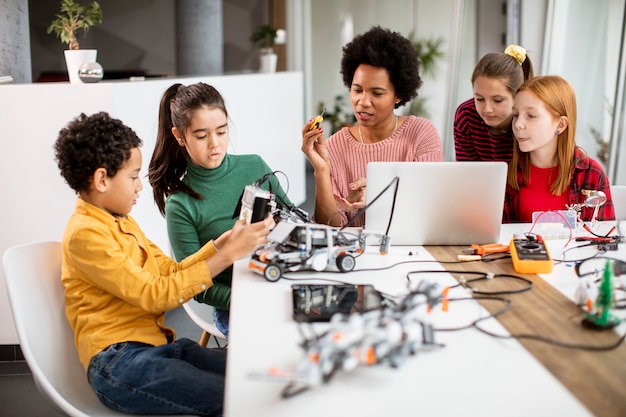 Groupe d'enfants heureux avec leur professeur de sciences afro-américaine avec des jouets électriques de programmation d'ordinateur portable et des robots en classe de robotique