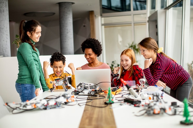 Photo groupe d'enfants heureux avec leur professeur de sciences afro-américaine avec des jouets électriques de programmation d'ordinateur portable et des robots en classe de robotique