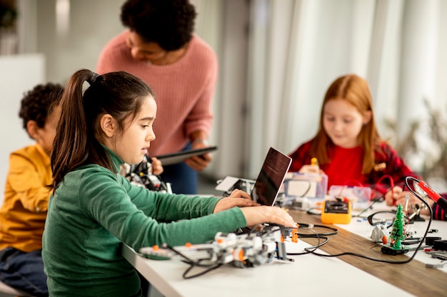 Groupe d'enfants heureux avec leur professeur de sciences afro-américaine avec des jouets électriques de programmation d'ordinateur portable et des robots en classe de robotique