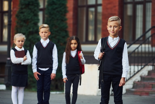 Photo groupe d'enfants en uniforme scolaire posant devant la caméra à l'extérieur ensemble près du bâtiment de l'éducation.