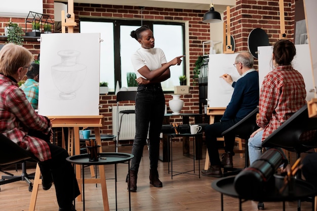 Photo groupe d'étudiants adultes apprenant à dessiner lors de cours d'art. une jeune enseignante afro-américaine amicale aide les gens pendant le cours de dessin, enseignant des techniques de dessin pour les débutants