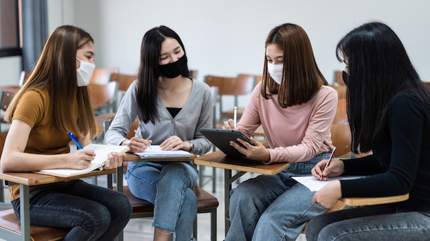 Groupe d'étudiants internationaux divers portant des masques de protection et parlant, discutant du projet, assis au bureau dans la salle de classe à l'université