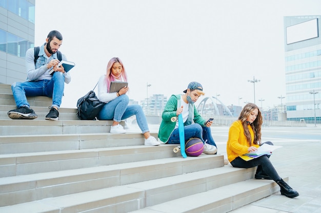 Un groupe d'étudiants multiraciaux portant des masques de protection étudient assis dans des escaliers à distance sociale devant un collège - Des amis heureux à l'heure du coronavirus font leurs devoirs à l'extérieur de l'école