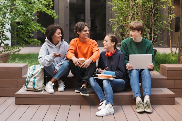 Groupe d'étudiants souriants cool assis et se regardant joyeusement tout en passant du temps ensemble dans la cour de l'université