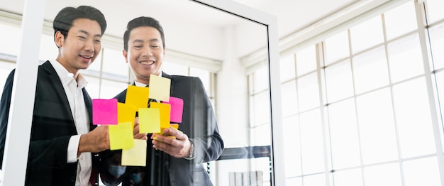 Groupe d'hommes d'affaires asiatiques prospères Travail d'équipe Réunion de remue-méninges avec des notes de papier collant sur le mur de verre pour de nouvelles idées Utilisation d'une méthodologie agile pour les entreprises dans un bureau de démarrage technologique