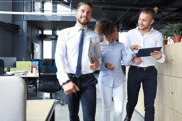 Un groupe d'hommes d'affaires modernes travaillent avec un ordinateur portable et sourient en se tenant debout dans le couloir du bureau.