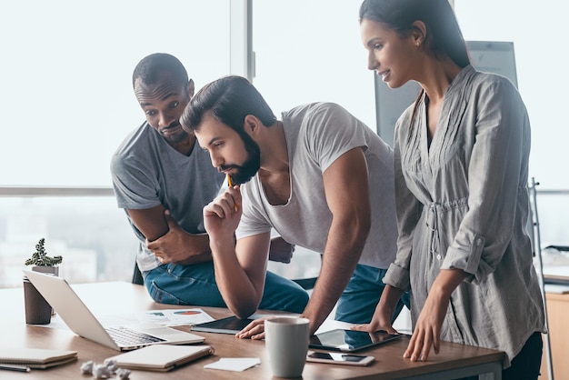 Groupe d'hommes d'affaires multiraciaux attrayants travaillant ensemble dans un bureau créatif tout en se tenant près du bureau en bois avec un ordinateur portable dessus, une équipe commerciale multiethnique amicale regardant sur l'écran, d