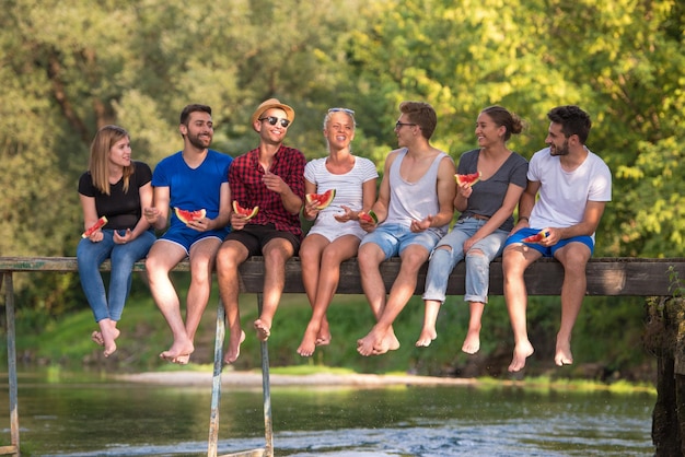 groupe de jeunes amis appréciant la pastèque tout en étant assis sur le pont en bois au-dessus de la rivière dans la belle nature