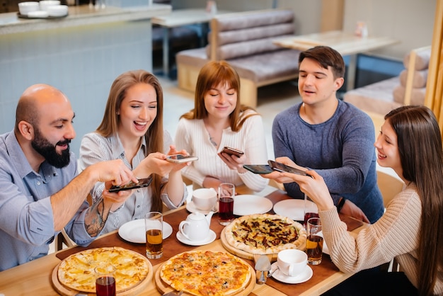 Un groupe de jeunes amis joyeux est assis dans un café en train de parler et de prendre des selfies au téléphone