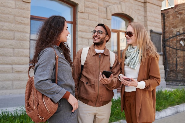 Groupe de jeunes amis multiethniques positifs qui vérifient leurs téléphones et discutent entre eux tout en passant du temps ensemble à l'extérieur