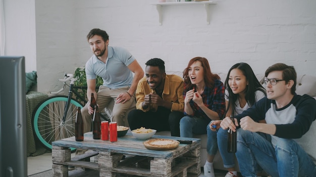 Groupe de jeunes amis qui regardent le match des jeux olympiques à la télévision ensemble en train de manger des collations et de boire de la bière Certains d'entre eux sont satisfaits de la victoire de leur équipe mais d'autres déçus