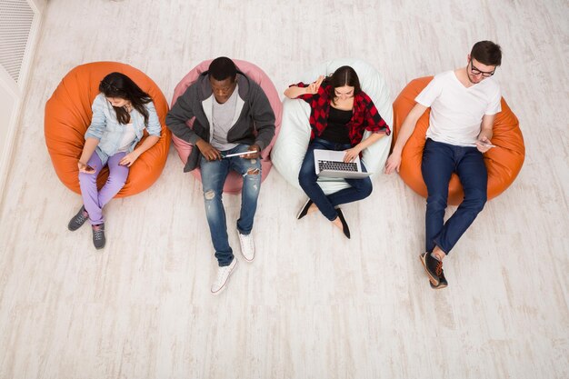 Groupe de jeunes amis utilisant des gadgets assis à l'intérieur du campus universitaire et travaillant ensemble sur une tâche créative, vue de dessus