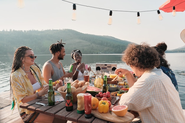 Groupe de jeunes assis à la table et se parler pendant le dîner de vacances sur une jetée