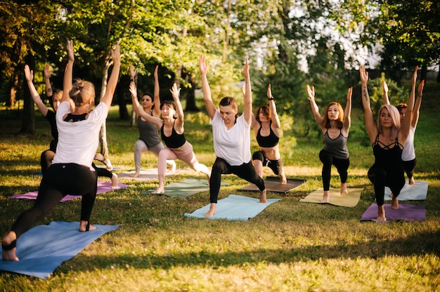 Groupe de jeunes belles femmes sportives pratiquant une leçon de yoga avec instructeur dans le parc de la ville le matin ensoleillé d'été. Un groupe de personnes se tient ensemble dans l'exercice Virabhadrasana 1, Warrior one pose