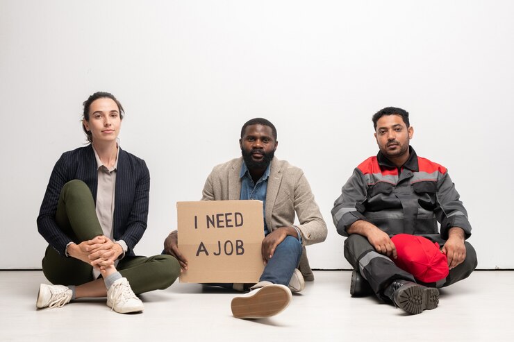 Photo groupe de jeunes chômeurs interculturels en tenue décontractée assis sur le sol devant la caméra, l'un d'eux tenant une note sur le besoin d'emploi