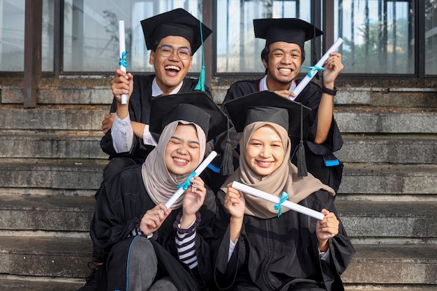 Groupe de jeunes étudiants divers dans des vêtements de graduation assis sur des escaliers avec des diplômes en mains looki