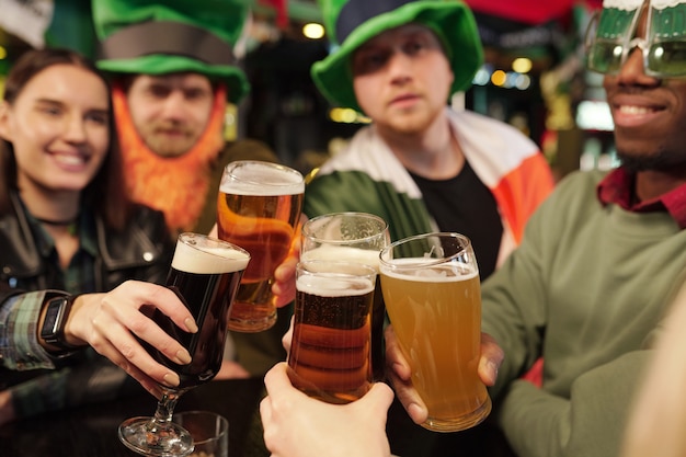 Groupe de jeunes fans de football heureux en tenue traditionnelle irlandaise tintant avec des verres de bière devant la caméra tout en célébrant la Saint-Patrick