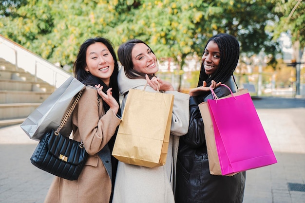 Un groupe de jeunes femmes diverses et heureuses marchant avec des sacs à provisions et se retournant en souriant pour regarder et saluer la caméra dans une rue urbaine Concept de consommation