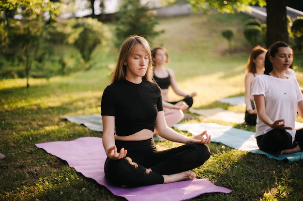 Un groupe de jeunes femmes est assise en position du lotus, les yeux fermés, faisant du yoga sur des tapis de yoga sur de l'herbe verte dans le parc le matin au lever du soleil.