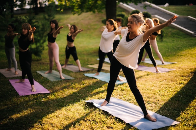 Groupe de jeunes femmes font du yoga pose dans le parc de la ville le matin ensoleillé d'été sous la direction d'un instructeur