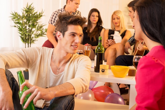 Un groupe de jeunes en fête à la maison. Au premier plan, un beau jeune homme assis par terre, parlant à une fille, et en arrière-plan un groupe de jeunes assis sur le canapé.