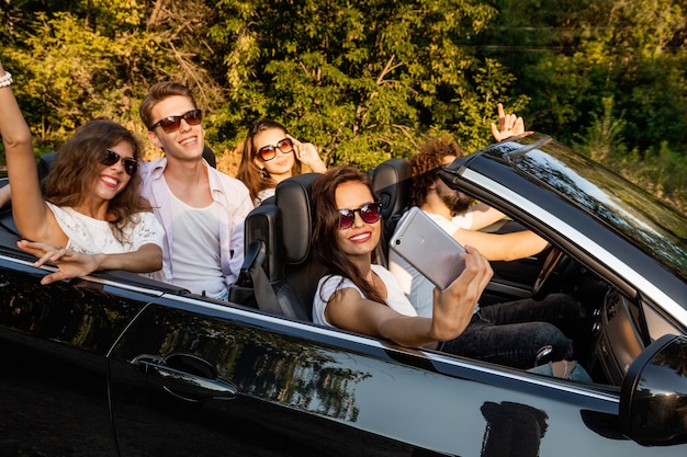 Un groupe de jeunes filles et de garçons font un selfie dans un cabriolet noir par une chaude journée ensoleillée. .