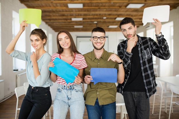 Groupe de jeunes gens joyeux tenant des icônes de messages de papiers colorés dans les mains regardant joyeusement à huis clos tout en passant du temps au travail dans un bureau moderne