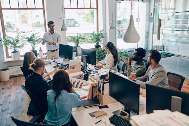 Groupe de jeunes gens modernes en vêtements décontractés intelligents communiquant et utilisant des technologies modernes tout en travaillant au bureau