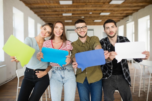 Groupe de jeunes gens souriants montrant des icônes de messages de papiers colorés dans les mains regardant joyeusement à huis clos tout en passant du temps au travail dans un bureau moderne