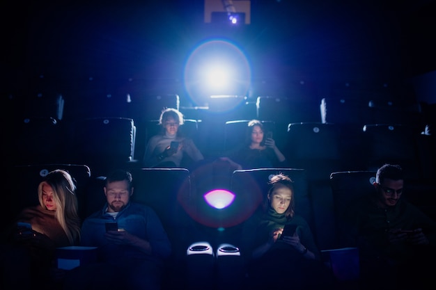Groupe de jeunes mobiles défilant dans leurs smartphones dans un cinéma sombre en attendant le début du film