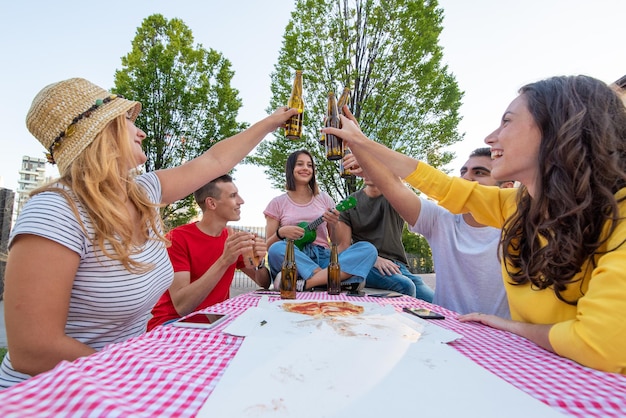 Groupe de jeunes portant un toast avec une bouteille de bière couples heureux d'amis s'amusant autour d'une table de pique-nique avec pizza et jouant des instruments de musique des millénaires insouciants chantant et souriant