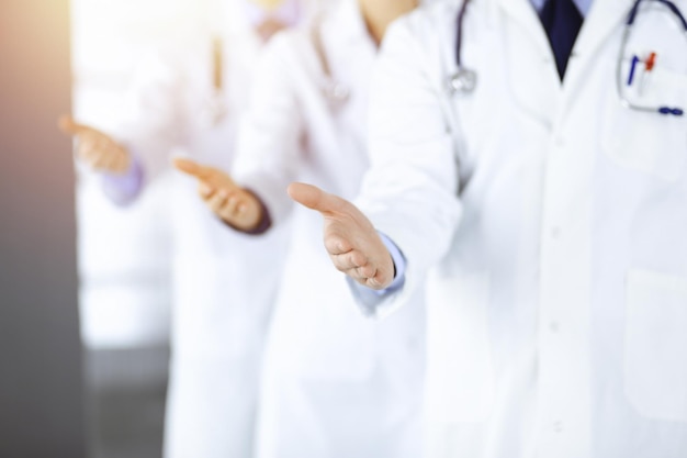 Un groupe de médecins modernes inconnus se tiennent comme une équipe avec les mains ouvertes dans un bureau d'hôpital ensoleillé. Des médecins prêts à examiner et à aider les patients. Aide médicale, assurance dans les soins de santé, meilleurs traitements contre les maladies